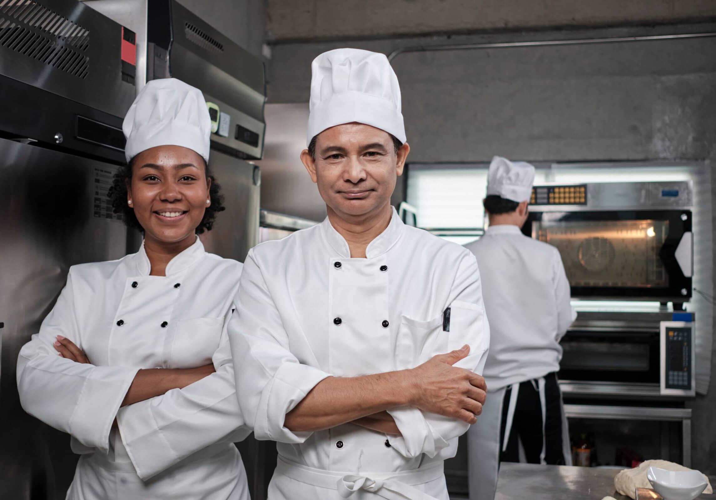 Pastry foods professional partners, two happy chefs team staff in white cooking uniforms stand, arms crossed with confidence, cheerful smiles with commercial culinary jobs in restaurant kitchen.